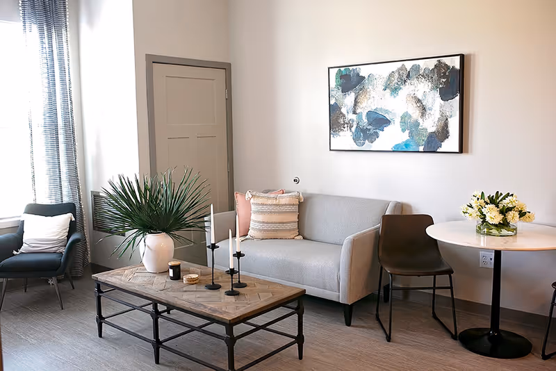 A modern living room with a gray sofa adorned with two pillows, a black chair, a wooden coffee table with decorative items including a white vase with green leaves and three black candlesticks, a round white table with a bouquet of white flowers, and a large abstract painting on the wall.