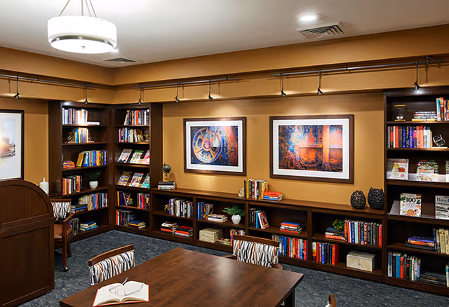 A cozy library reading area featuring bookshelves filled with various books, a wooden table with an open book, and framed artwork on the walls.