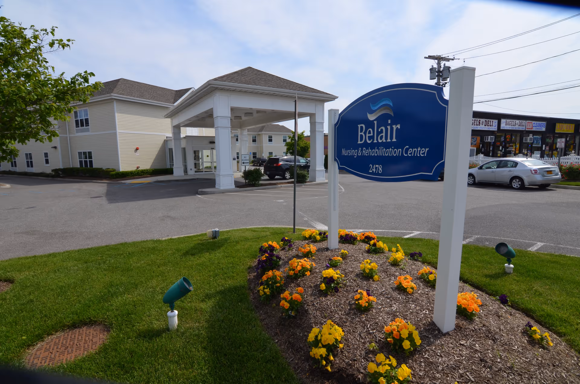 Exterior view of Belair Nursing & Rehabilitation Center showing the entrance with a covered drop-off area, a blue sign with the facility name and address, a flower bed with yellow and orange flowers, a parking lot with cars, and a strip mall in the background.