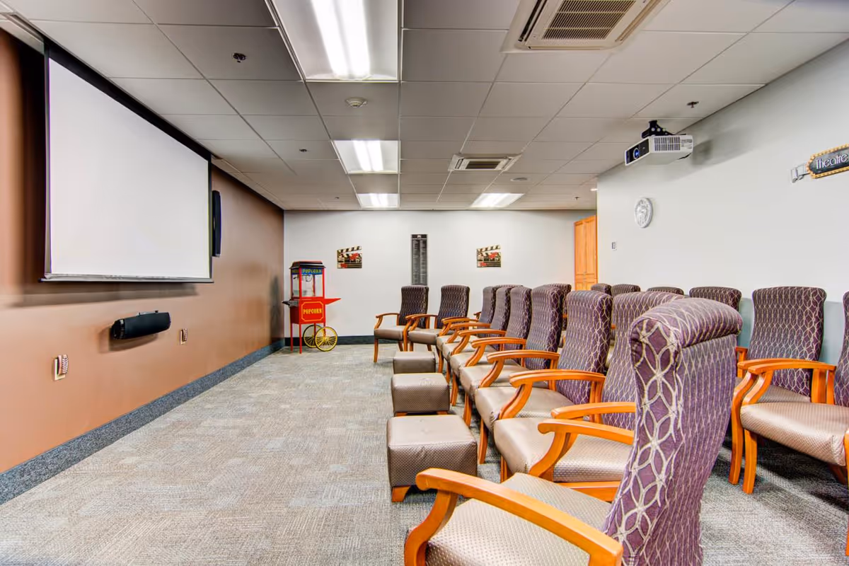 A small theater room with rows of upholstered chairs with wooden armrests facing a large white projection screen on a brown wall. There is a popcorn machine in the back corner and a projector mounted on the ceiling. The room has a carpeted floor and fluorescent ceiling lights.