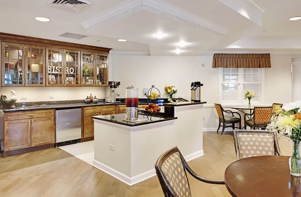 A bright and clean kitchen and dining area in a senior living facility. The kitchen features wooden cabinets with glass doors, a countertop with a beverage dispenser and coffee pots, and a small island. The dining area has round tables with cushioned chairs and floral centerpieces, with a window covered by a striped valance.