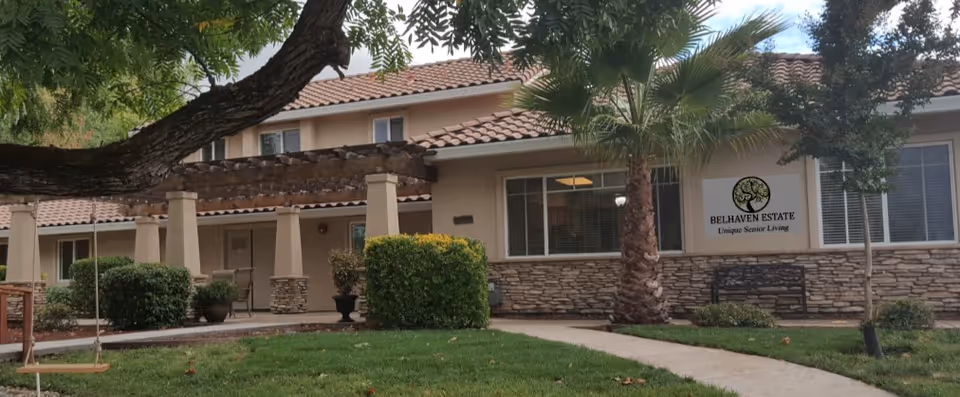 Exterior view of Belhaven Estate senior living facility showing a two-story building with a tiled roof, stone facade, and a covered porch area. There is a green lawn with a tree swing in the foreground and several bushes and palm trees near the building.