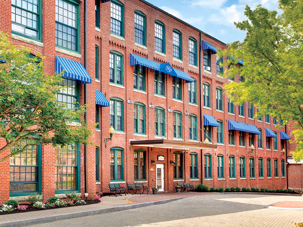Exterior view of a red brick building with green window frames and blue striped awnings. There are benches and flower beds along the sidewalk, and trees with green leaves on either side of the building.
