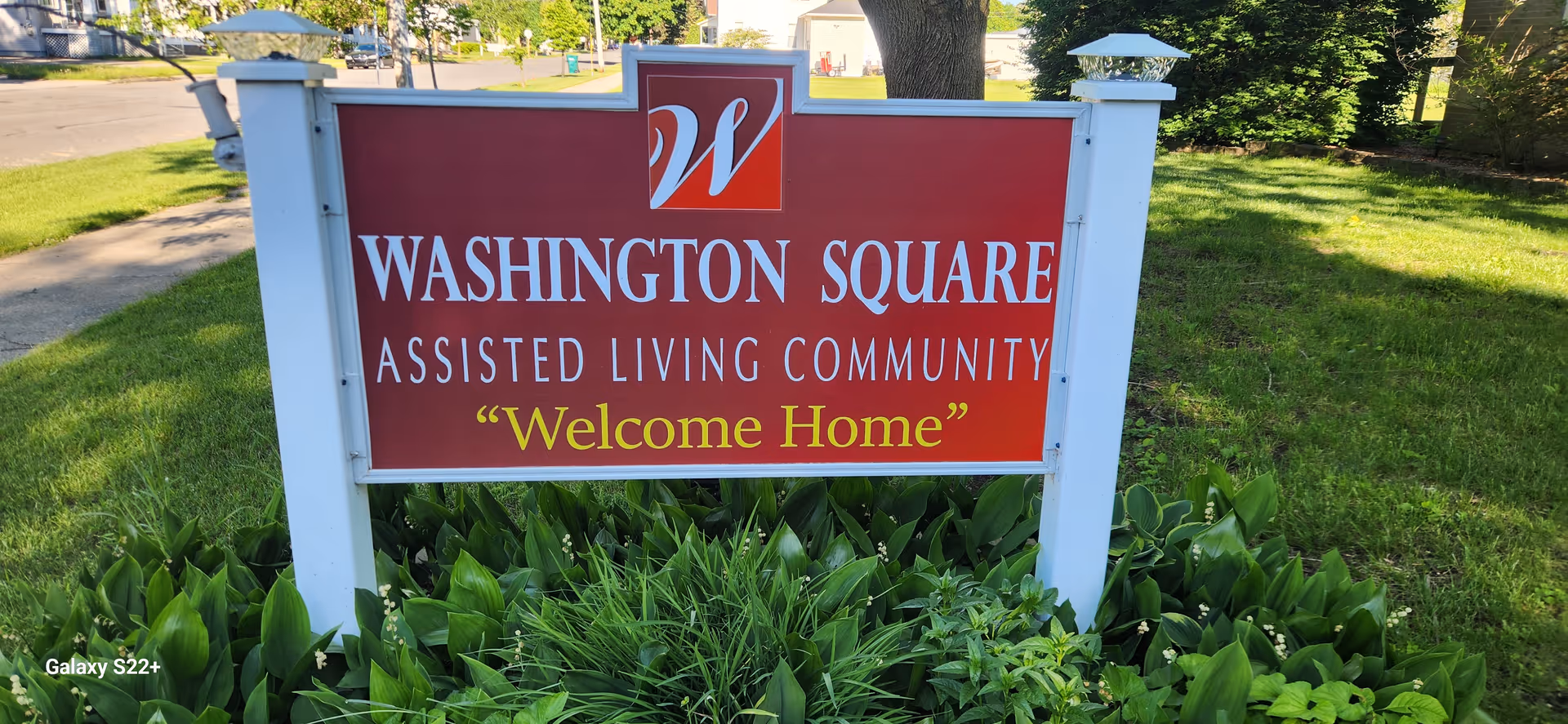 A red and white sign for Washington Square Assisted Living Community with the slogan 'Welcome Home' displayed in yellow text. The sign is mounted on white posts and surrounded by green plants and grass, with a sidewalk and street visible in the background.