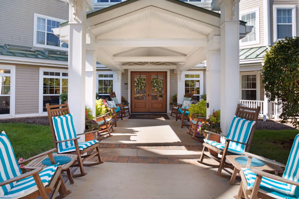 Covered entrance area of a senior living facility with wooden rocking chairs featuring blue and white striped cushions arranged on both sides. The entrance has double wooden doors decorated with wreaths, surrounded by windows and potted plants.