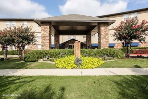 Front exterior view of a senior living facility named Morada Meadow Lakes featuring a covered entrance with brick pillars, well-maintained landscaping including bushes and flowering trees, and a clear blue sky with some clouds.