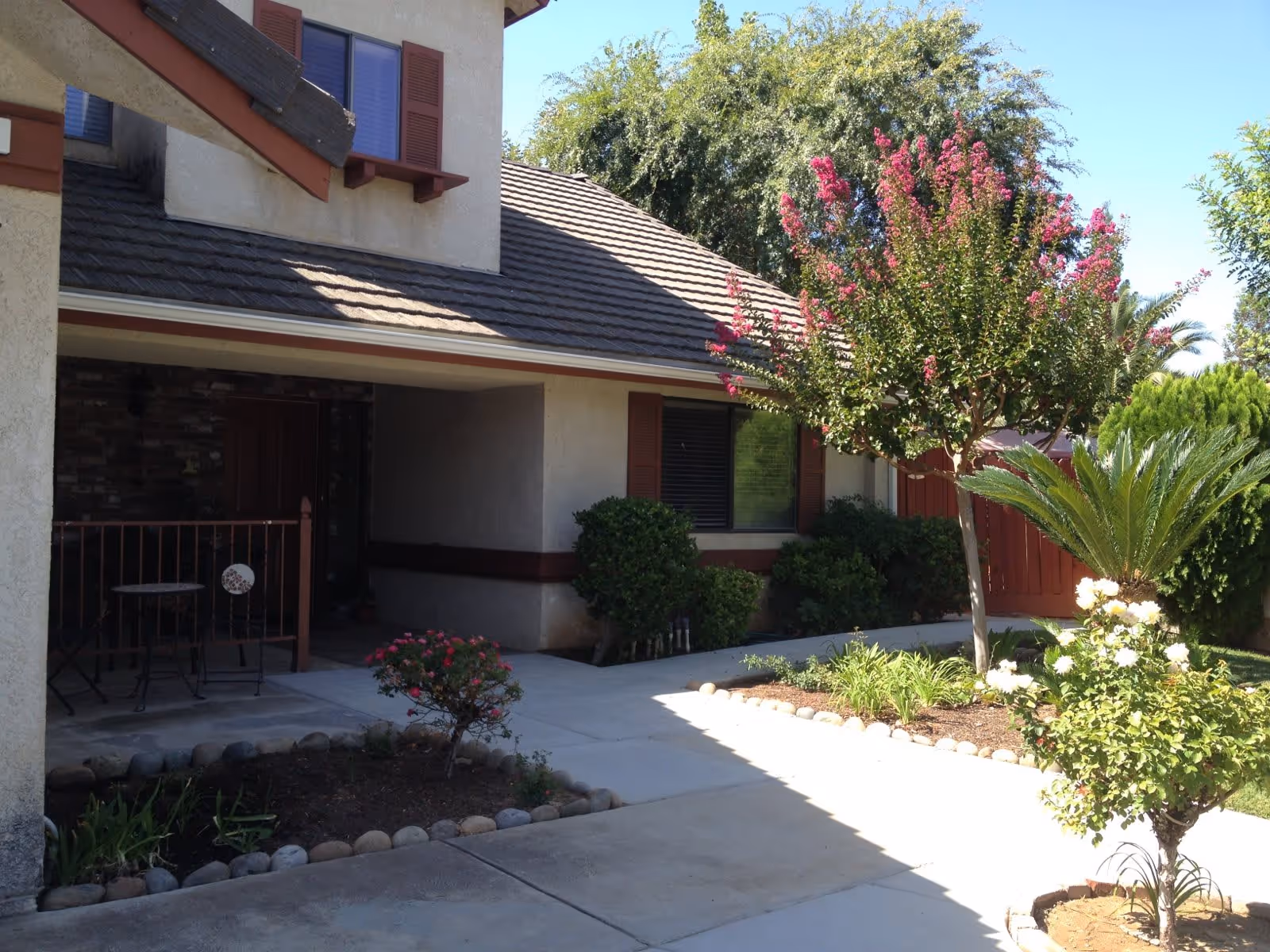Outdoor view of a residential facility showing a walkway leading to a covered porch area with a small table and chairs. The building has beige walls with brown trim and a tiled roof. There are various plants, bushes, and a tree with pink flowers in landscaped garden beds along the walkway.