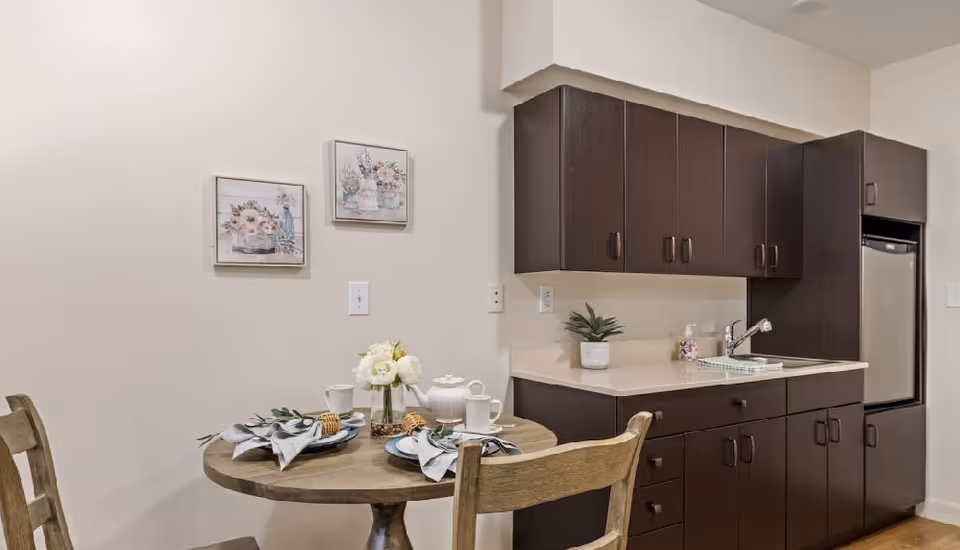 A small dining area with a round wooden table set for two with plates, cups, napkins, and a vase of white flowers. Behind the table is a kitchenette with dark brown cabinets, a countertop with a small plant, a sink, and a compact refrigerator. Two framed floral pictures hang on the beige wall above the table.