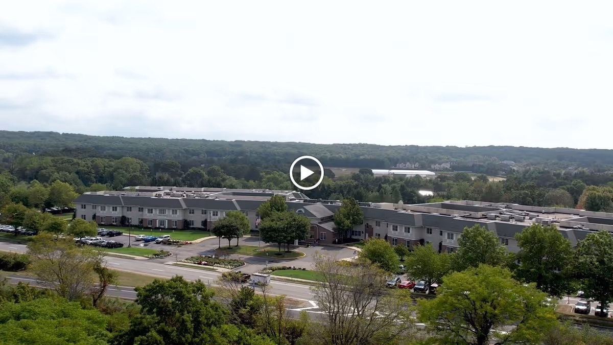 Aerial view of Huntington Hills Center for Health and Rehabilitation, a large two-story building surrounded by trees and parking lots, with a road in the foreground and a forested area in the background under a cloudy sky.