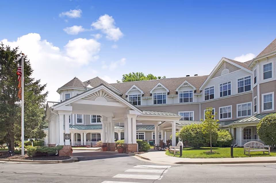 Exterior view of a senior living facility building with a covered entrance, multiple windows, and a well-maintained garden area with benches and trees under a blue sky with some clouds.