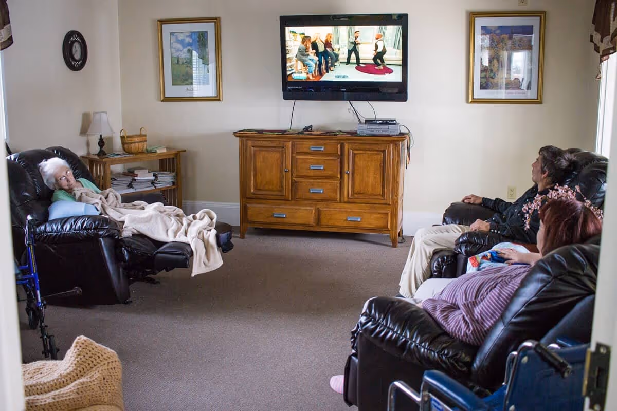 A cozy living room with three elderly people sitting on black leather recliners watching a wall-mounted TV. The room has beige walls, a wooden cabinet under the TV, framed pictures on the walls, a small side table with a lamp and books, and a walker near one recliner.