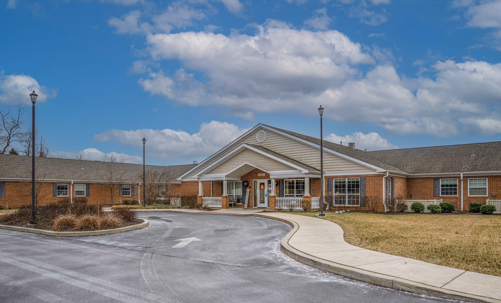 Exterior view of Northampton Post Acute facility showing a single-story brick building with a covered entrance, a curved driveway, lamp posts, and a partly cloudy blue sky.