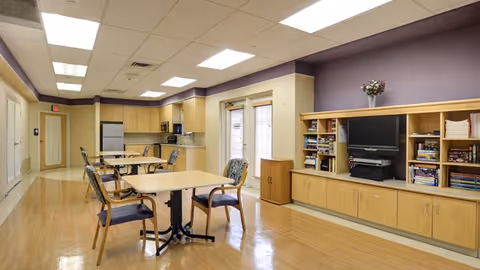 A common area in a senior living facility with light wood flooring, several tables and chairs, a kitchenette with a refrigerator and microwave, and a built-in entertainment center with a TV and shelves filled with books and games. The walls are painted in light beige and purple tones, and there are double doors with windows letting in natural light.