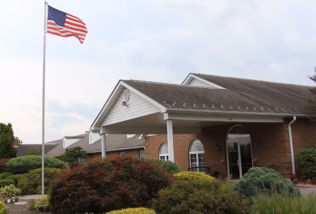 Exterior view of a brick building with a covered entrance and an American flag on a flagpole in front. The building is surrounded by well-maintained bushes and greenery under a cloudy sky.