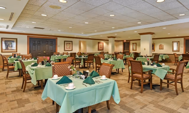 A spacious dining room with multiple tables covered in green tablecloths, each set with green folded napkins, white cups, and silverware. The room features patterned chairs, framed artwork on the walls, and a beige carpeted floor under a ceiling with recessed lighting.
