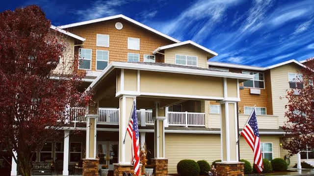 Exterior view of a multi-story senior living facility building with beige and brown siding, white railings on balconies, and two American flags displayed near the entrance under a covered porch. Trees with red leaves are visible on both sides and a partly cloudy blue sky is in the background.