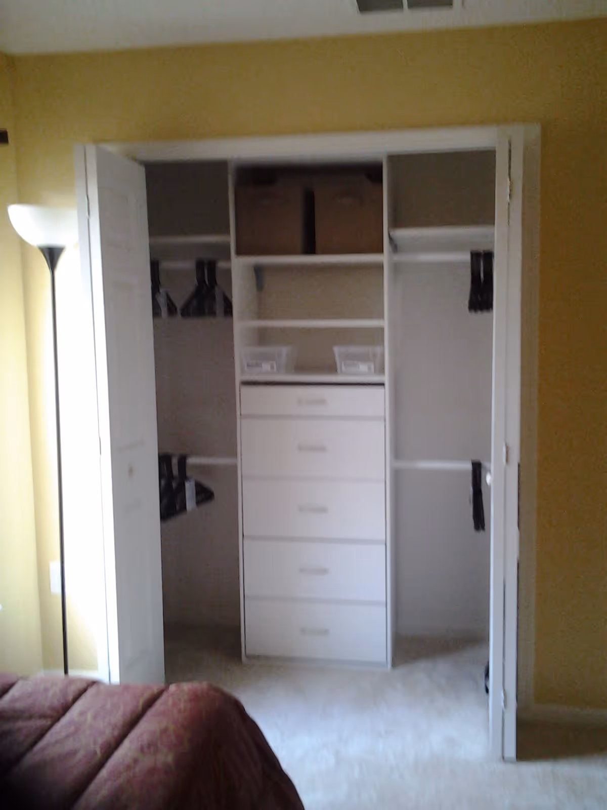 An open closet with white shelves and drawers inside a room with yellow walls. The closet contains several black hangers and two brown storage boxes on the top shelf. A floor lamp is visible to the left of the closet, and part of a red bedspread is visible in the foreground.