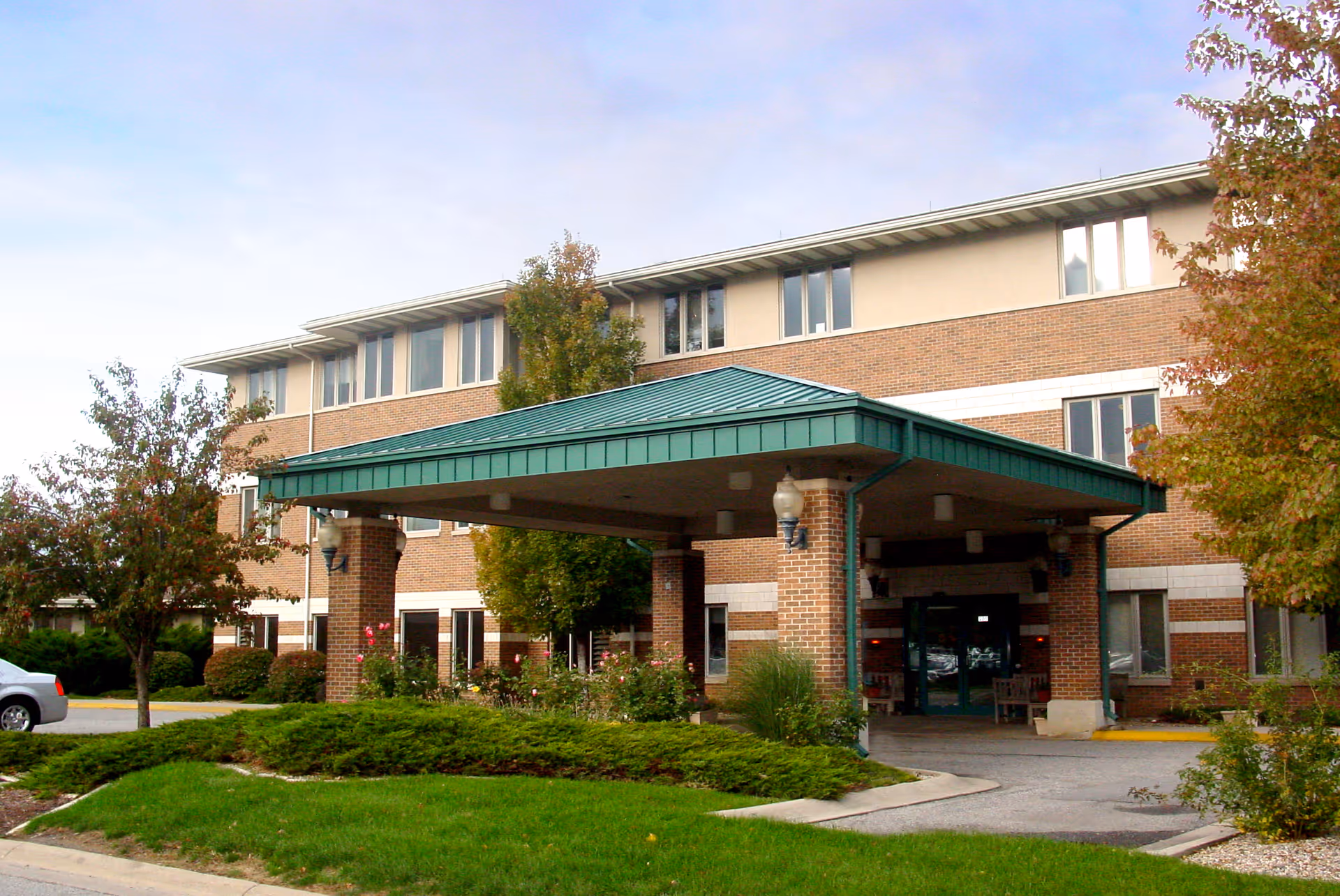 Exterior view of a three-story brick building with a green metal roof over the entrance. There are trees and shrubs around the entrance area, and a car is partially visible on the left side.