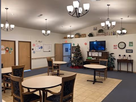A common area in a senior living facility with round tables and chairs arranged on a tiled and carpeted floor. The room is decorated with a Christmas tree, a television on a wooden stand, wall clocks, and various wall decorations. Multiple ceiling light fixtures illuminate the space.