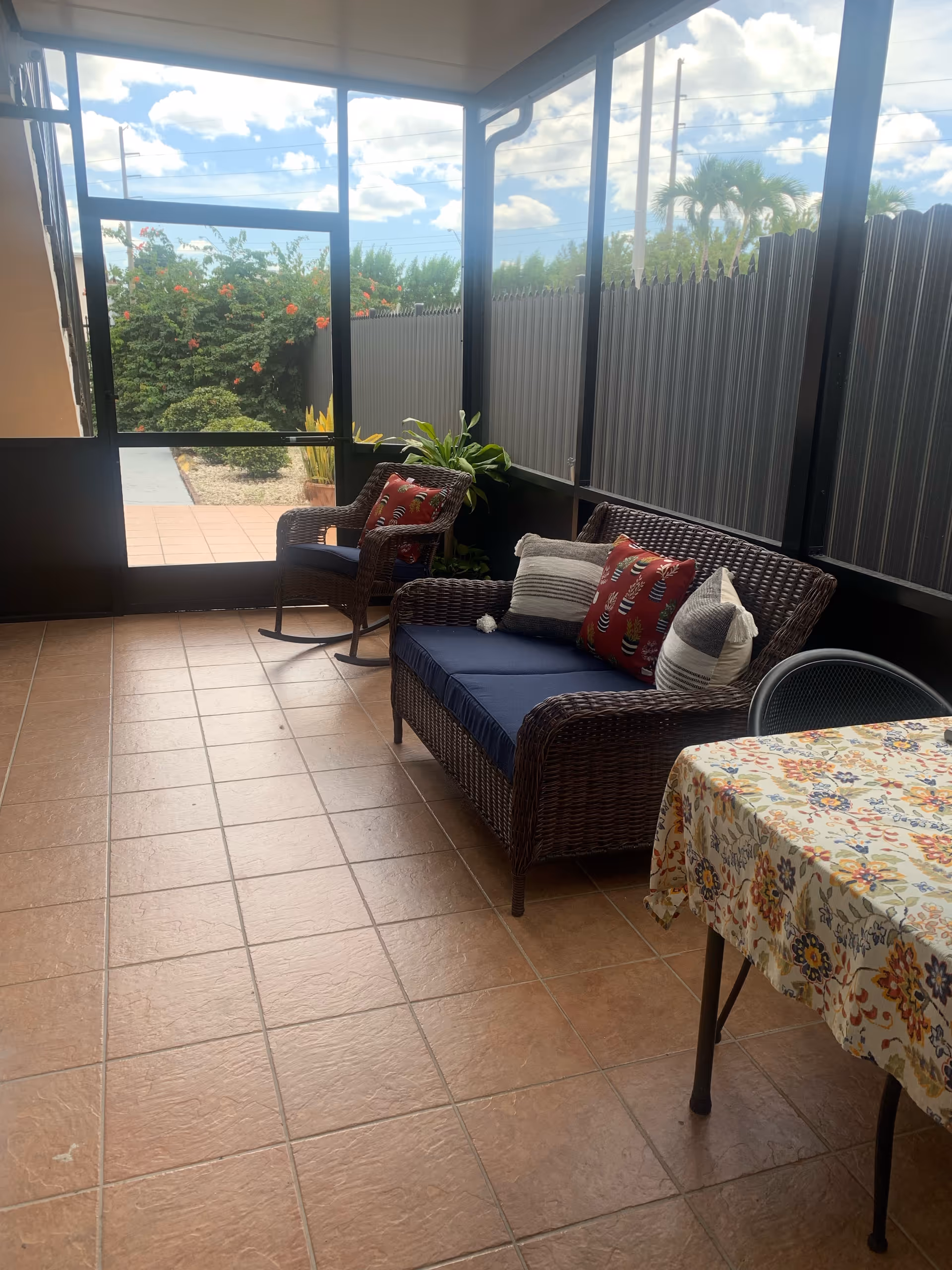 A screened-in porch area with tiled floor, featuring a wicker loveseat with blue cushions and decorative pillows, a matching wicker rocking chair with a cushion, a small plant, and a table covered with a floral tablecloth. Outside the screened area, there are bushes and a clear sky with clouds.