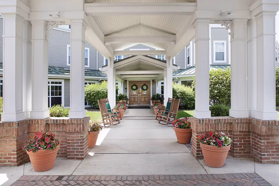 Covered entrance walkway to a building with white columns and brick bases, lined with potted plants and wooden rocking chairs, leading to double wooden doors decorated with wreaths. The building exterior is light gray with white trim and green metal roofing.