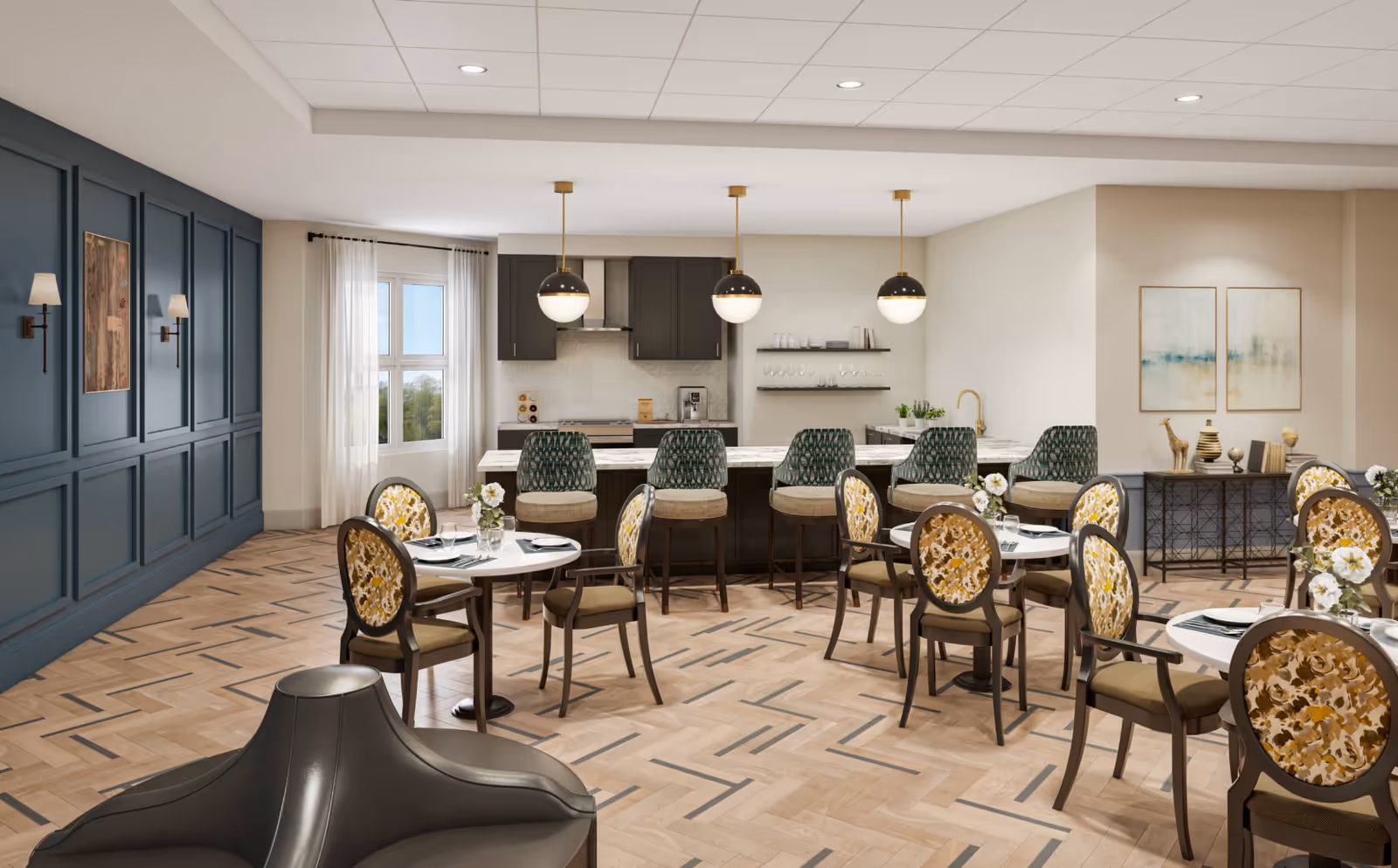 A spacious dining area with several round tables and chairs featuring floral upholstery. In the background, there is a kitchen area with a long marble countertop island and five bar stools with patterned backs. The kitchen has dark cabinets, three pendant lights hanging above the island, and shelves with glassware. The room has a large window with sheer white curtains, blue paneled walls with sconces, and light wood herringbone flooring.