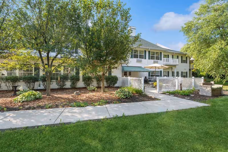 Exterior view of a two-story residential building with white siding and green shutters, surrounded by trees and landscaped garden beds. A concrete walkway leads to a fenced patio area with outdoor seating and umbrellas under a clear blue sky.
