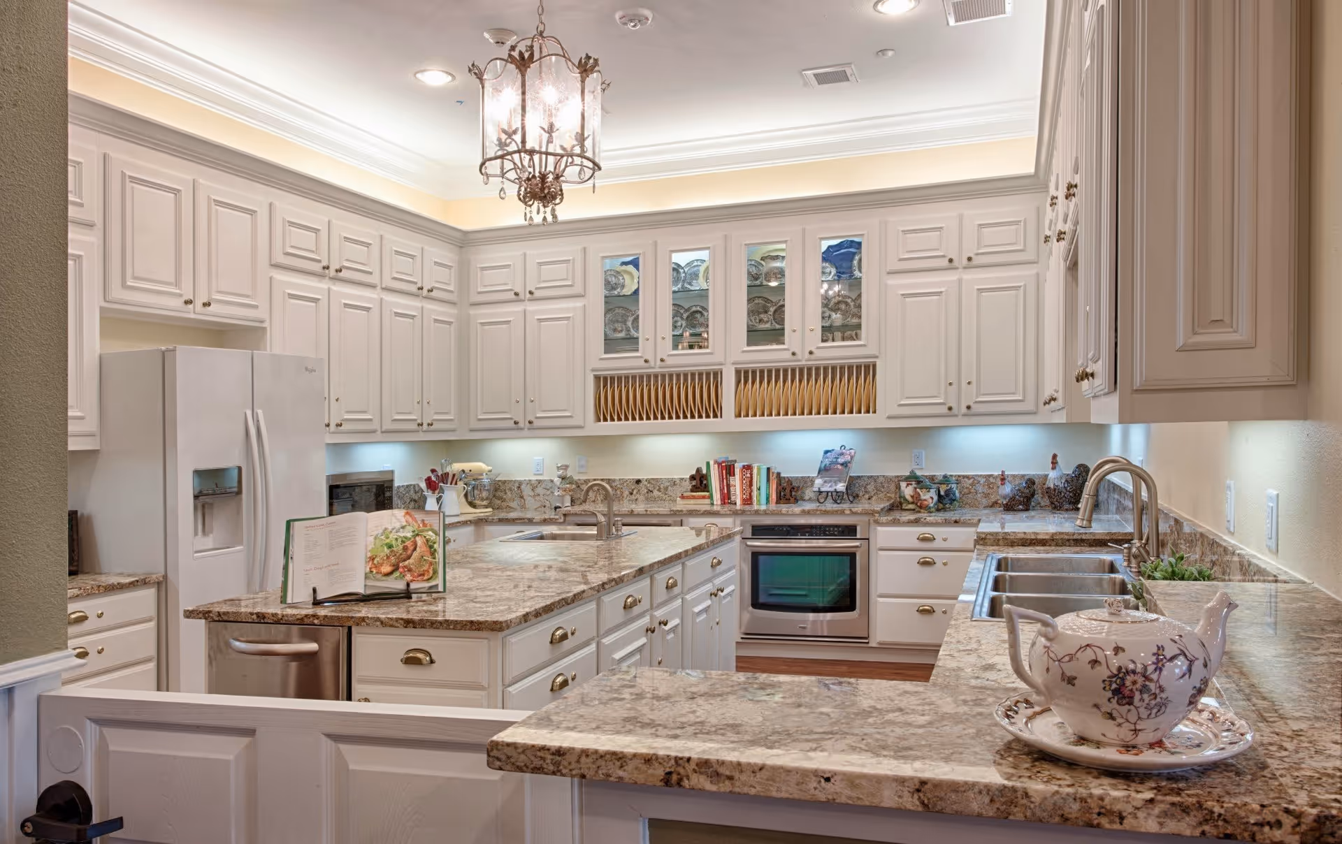 A spacious kitchen with white cabinetry and granite countertops. The kitchen features a large island with a built-in sink, a refrigerator, an oven, and a dishwasher. There is a decorative chandelier hanging from the ceiling and under-cabinet lighting illuminating the countertops. A floral teapot and an open cookbook are placed on the countertops.