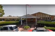 Front entrance of Berkshire Nursing & Rehabilitation Center with a covered drop-off area, an American flag on a flagpole, and several parked cars in front. The building is a single-story structure with large windows and a brick facade.