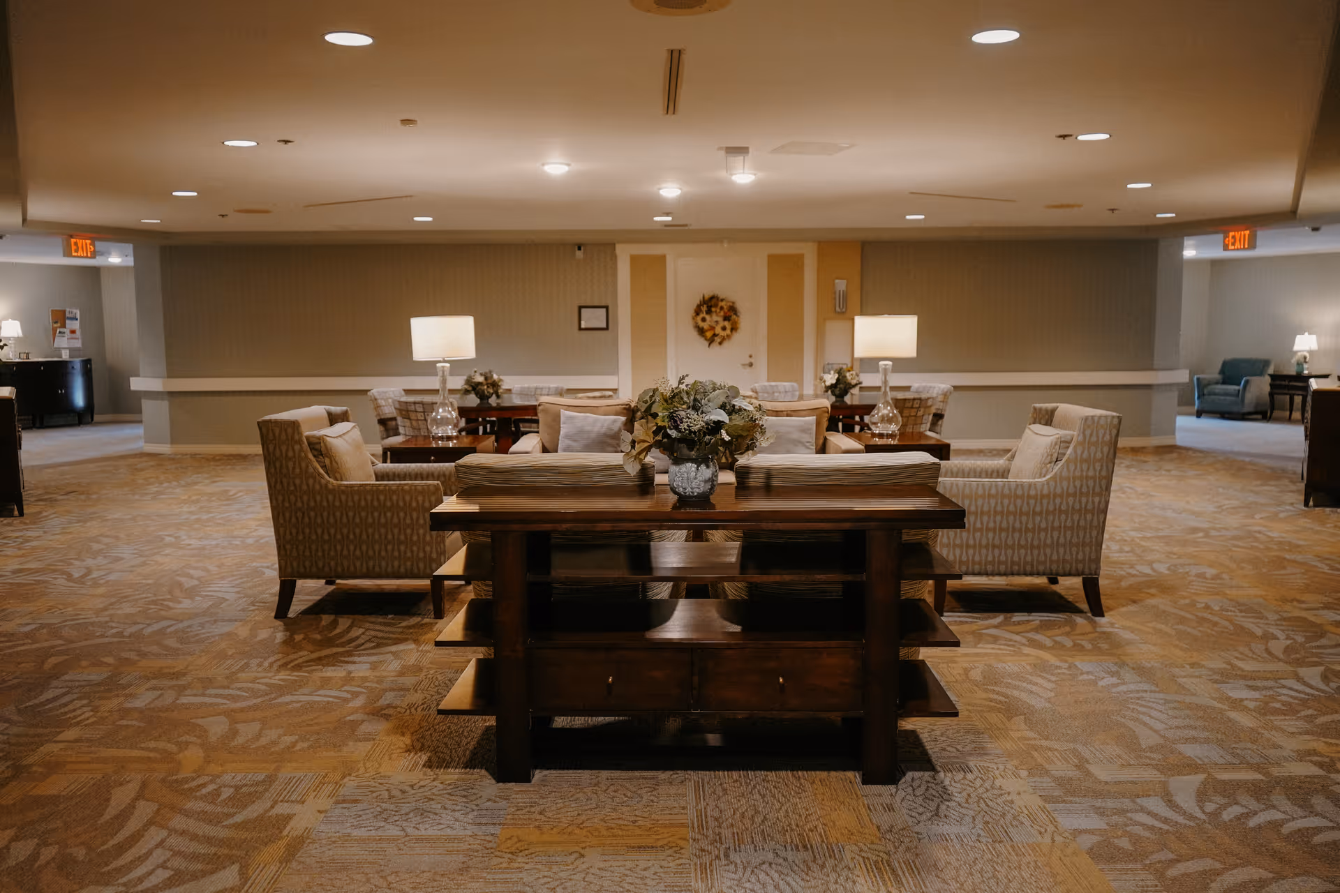 A spacious and well-lit common area with patterned carpet flooring, several armchairs arranged around wooden tables with lamps and floral centerpieces, and a wooden console table with a flower arrangement in the foreground. The walls are painted in neutral tones, and there are exit signs visible in the background.