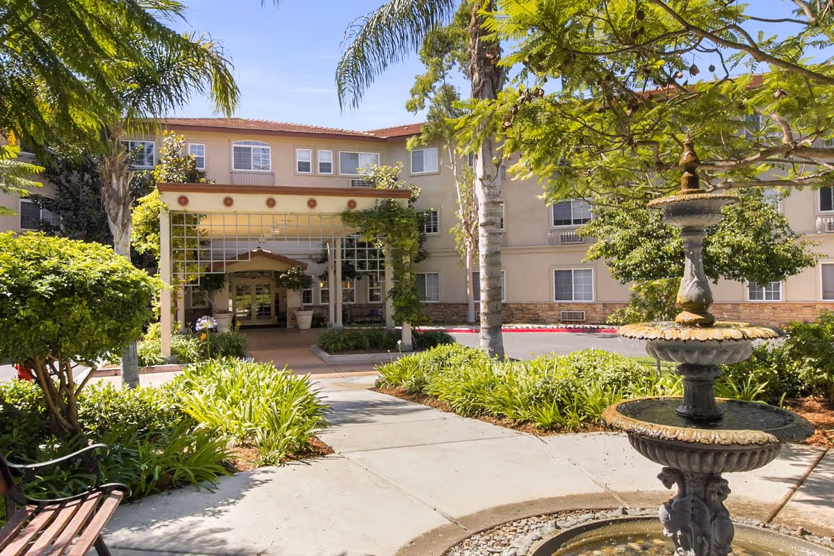 Exterior view of a senior living facility with a three-story building, surrounded by lush greenery, palm trees, and a decorative three-tiered fountain in the foreground. There is a paved walkway leading to the entrance, which has a covered porch with climbing plants and large potted plants on either side.