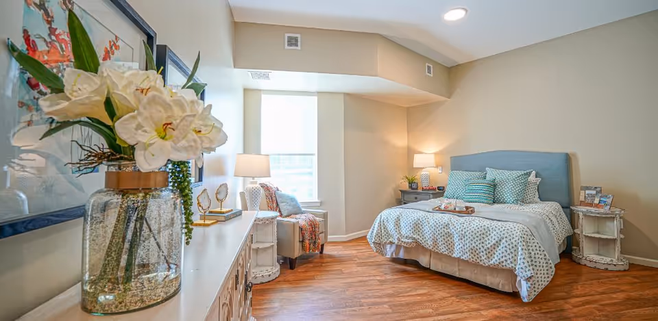 A bright and cozy bedroom with a large bed featuring a blue upholstered headboard and patterned bedding. There are multiple pillows on the bed and a tray with cups placed on it. To the right of the bed is a small round white nightstand with decorative items and a lamp. On the left side of the room, there is a window with a chair and a side table with a lamp. In the foreground, a large glass vase with white flowers is placed on a white dresser. The room has wooden flooring and beige walls with recessed lighting.
