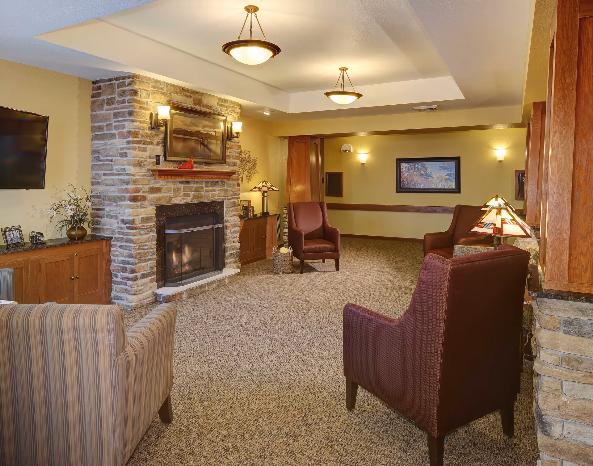 A cozy living room area in Heritage Assisted Living & Memory Care featuring a stone fireplace with a framed picture above it, surrounded by wooden cabinets. The room has several comfortable armchairs in burgundy and striped upholstery, warm lighting from ceiling fixtures and lamps, and a carpeted floor.