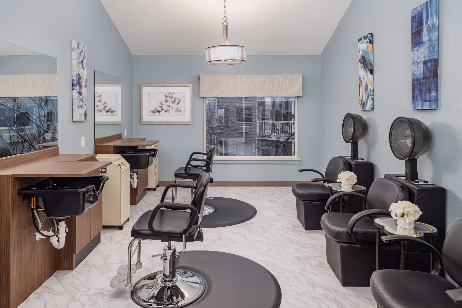 Interior view of a salon area with two black salon chairs in front of sinks and mirrors on the left side, and three black chairs with hair dryers on the right side. The walls are painted light blue with abstract artwork, and there is a window with a beige valance at the back.