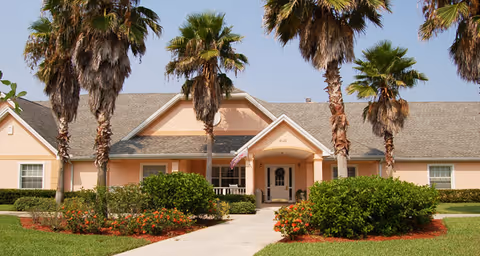 Front exterior view of a single-story building with a peach-colored facade, a gray shingled roof, and several tall palm trees in front. There is a concrete walkway leading to the entrance, which has a small covered porch with an American flag hanging. The landscaping includes green bushes and flowering plants.