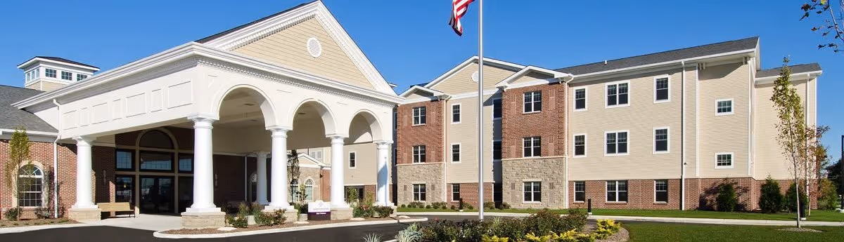 Exterior view of St Clare Commons senior living facility showing a three-story building with beige and brick facade, a covered entrance with white columns, an American flag on a flagpole, and landscaped grounds under a clear blue sky.
