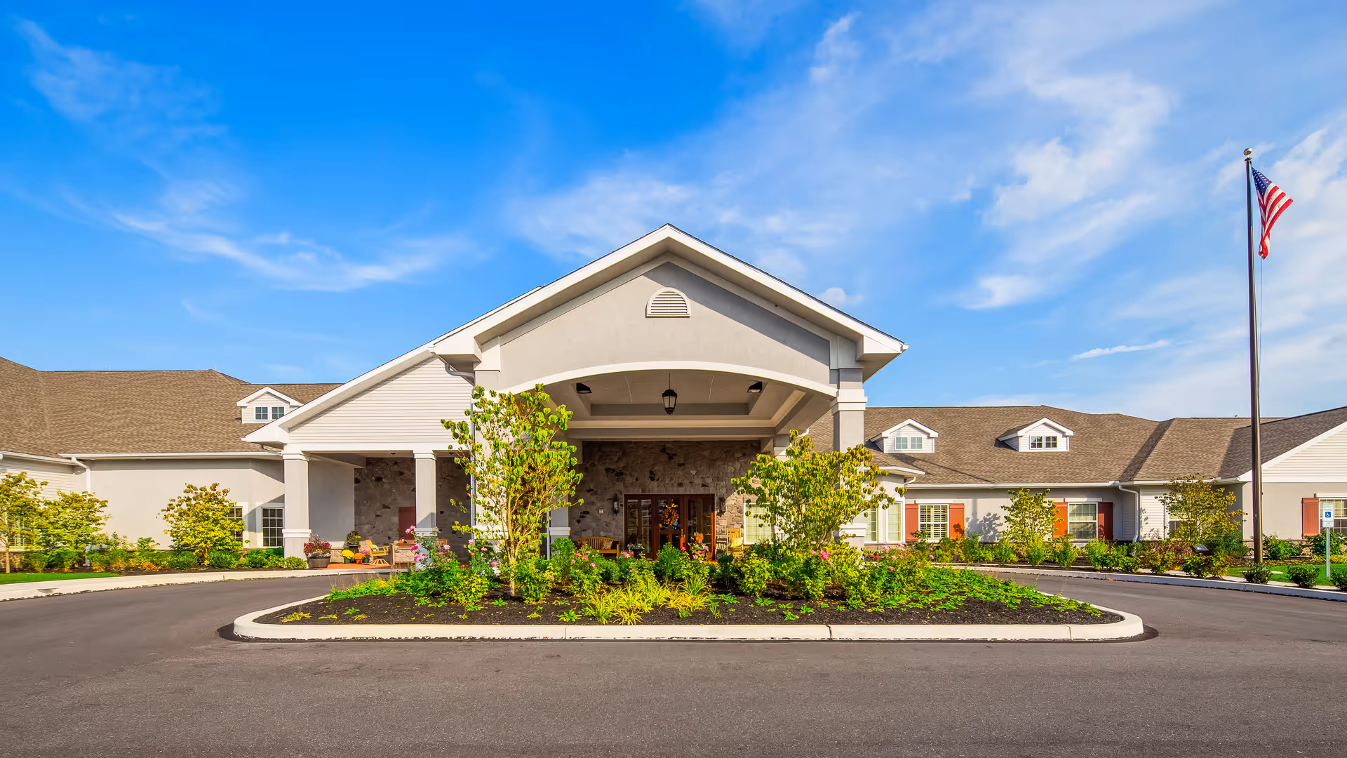 Front exterior view of the Legend of Lancaster senior living facility with a covered entrance, landscaped greenery, and an American flag on a flagpole under a blue sky.