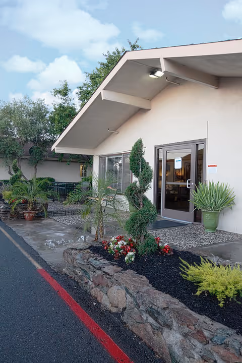 Exterior view of a single-story building with a slanted roof, featuring a glass door entrance and windows. The entrance is surrounded by landscaped plants, including a spiral-shaped topiary, small palm trees, and colorful flowers. The sky is partly cloudy.