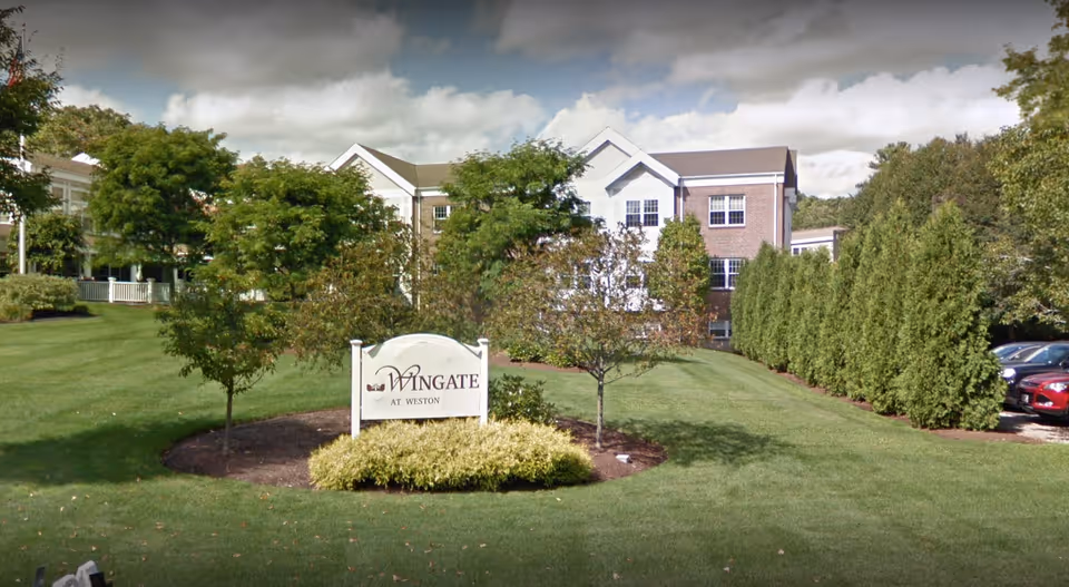 Exterior view of Wingate Residences at Weston, showing a landscaped lawn with trees, shrubs, and a sign with the facility name in front of a multi-story brick and white building under a partly cloudy sky.
