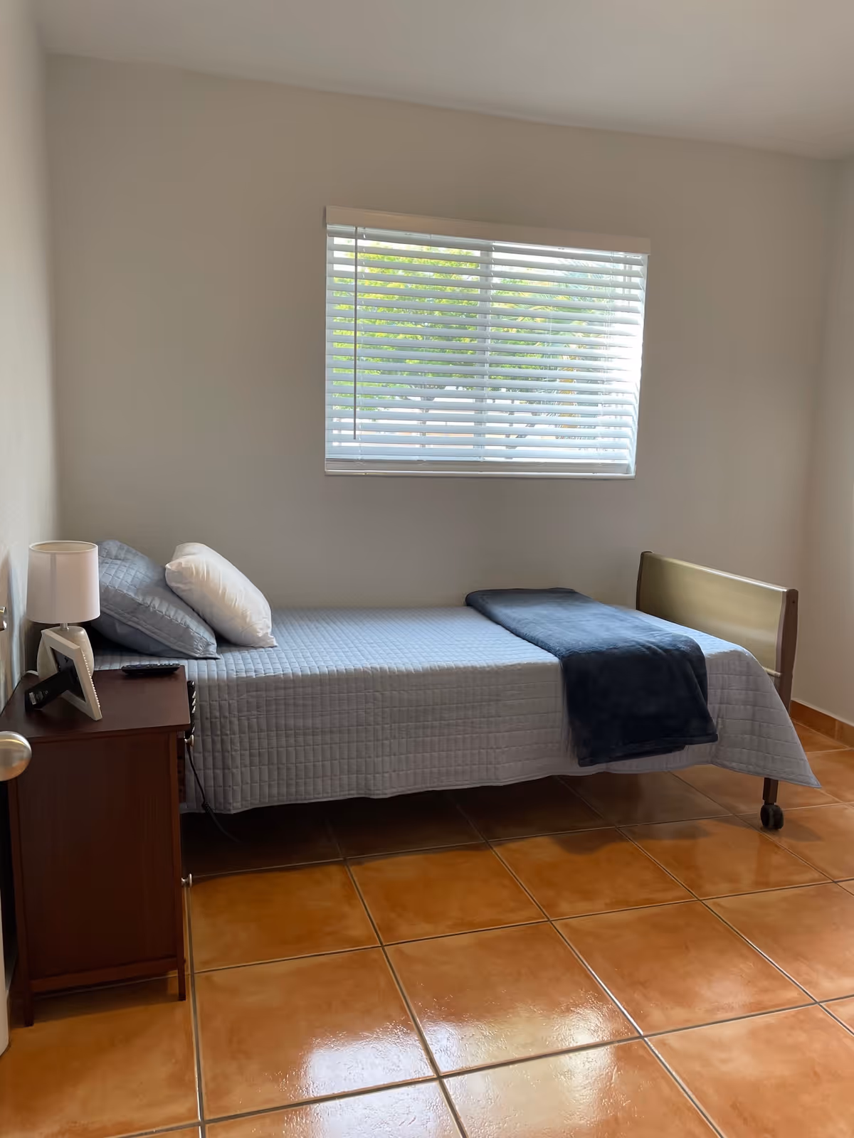 A simple bedroom with a single bed covered in a light blue quilt and a dark blue blanket folded at the foot. There are two pillows on the bed. Next to the bed is a wooden nightstand with a white lamp, a picture frame, and a remote control. A window with white blinds is on the wall above the bed, letting in natural light. The floor is covered with large, glossy, brown tiles.