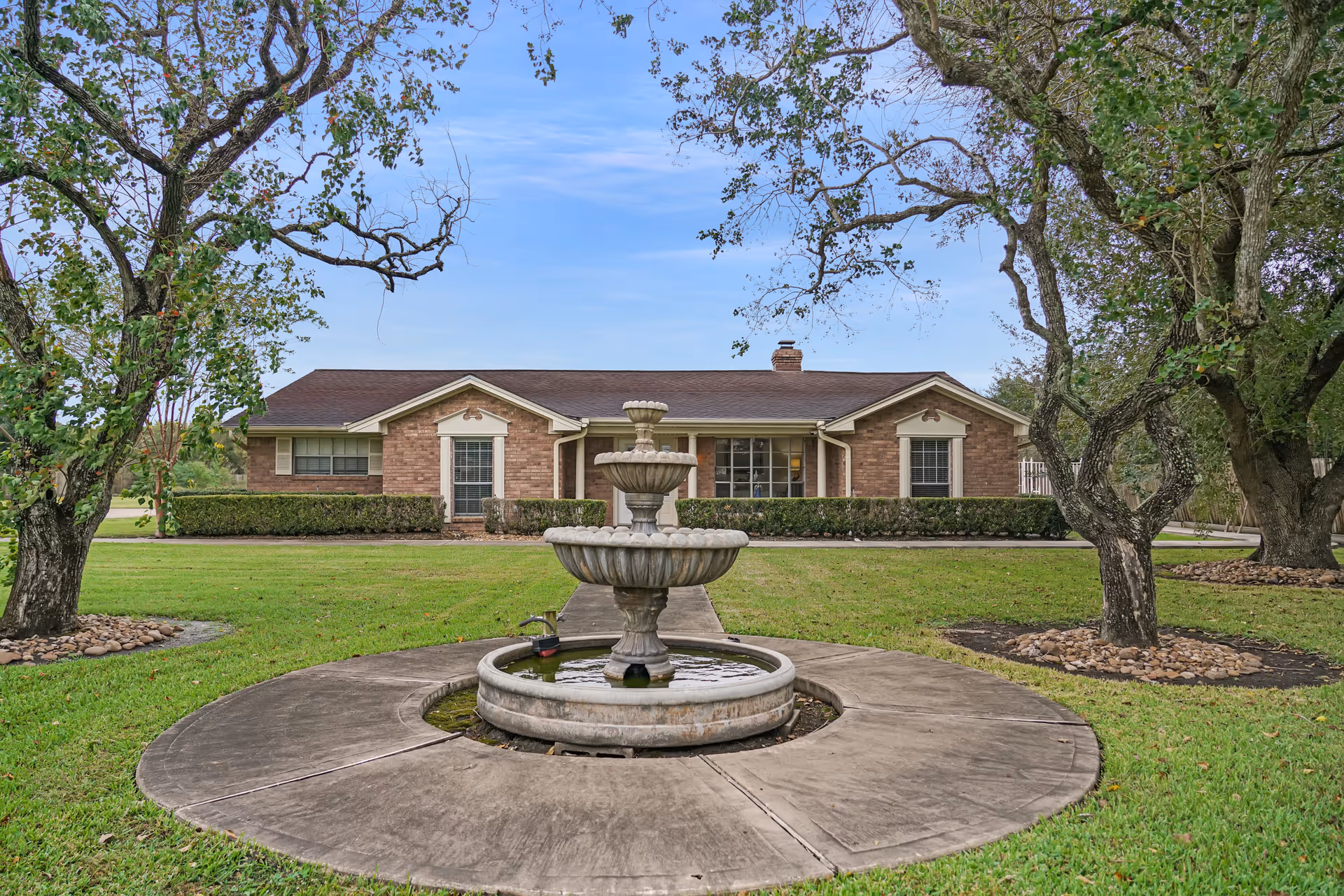 A single-story brick building with a brown roof, surrounded by green grass and trees. In the foreground, there is a circular concrete pathway around a tiered water fountain. The sky is clear with a few clouds.