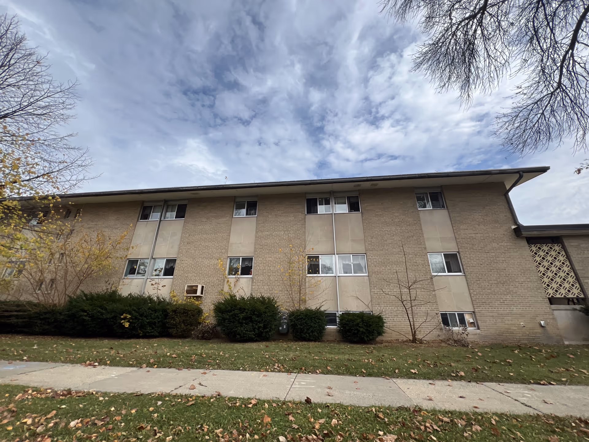 Exterior view of a two-story brick building with multiple windows, surrounded by bushes and trees with some leaves fallen on the grass and sidewalk in front. The sky is partly cloudy.