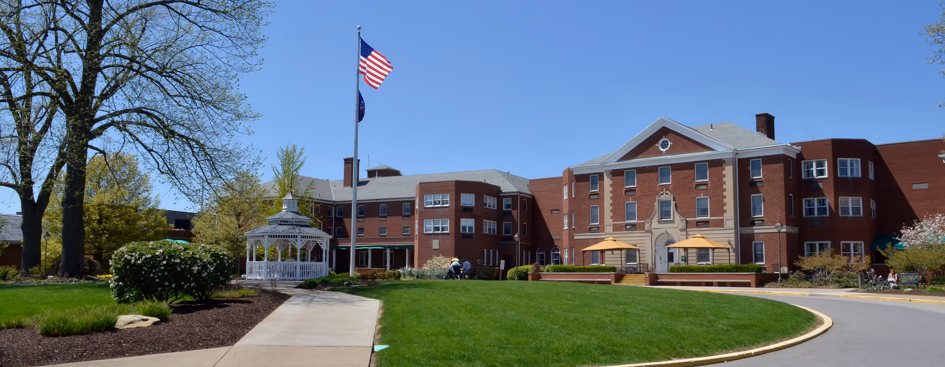 Exterior view of a large brick building with multiple windows and a gray roof, surrounded by green lawns, trees, and bushes. A white gazebo and two flagpoles with American and state flags are visible near the center. The sky is clear and blue.