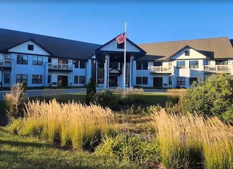 Exterior view of a two-story senior living facility building with white siding and dark roof. The building has balconies and large windows. In front, there is a landscaped area with tall grasses and shrubs, and an American flag on a flagpole. The sky is clear and blue.