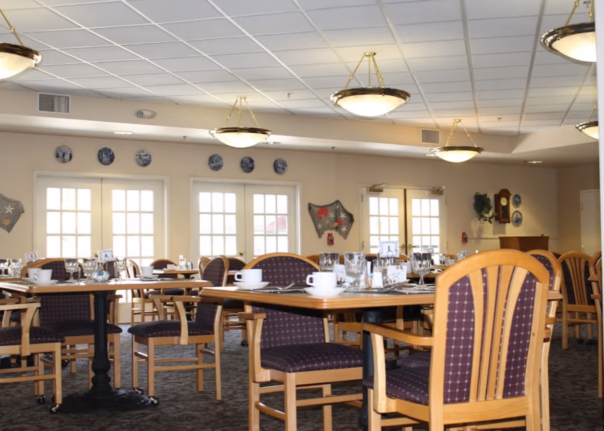 A dining room in a senior living facility with multiple wooden tables and chairs with purple cushioned seats. The tables are set with white cups, glasses, and silverware. The room has large windows and glass doors letting in natural light, with decorative plates and wall hangings on the walls. Ceiling lights hang from the white tiled ceiling.