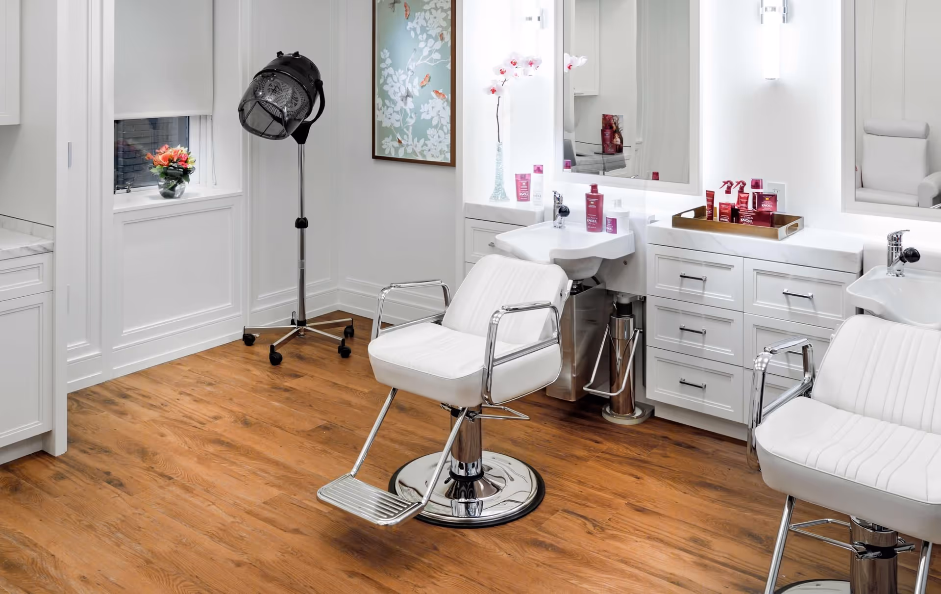 A bright and clean salon area with two white salon chairs, a hair dryer on a stand, a sink for washing hair, and a countertop with various hair care products. The room has wooden flooring, white walls, a window with a flower vase on the sill, and a framed floral artwork on the wall.