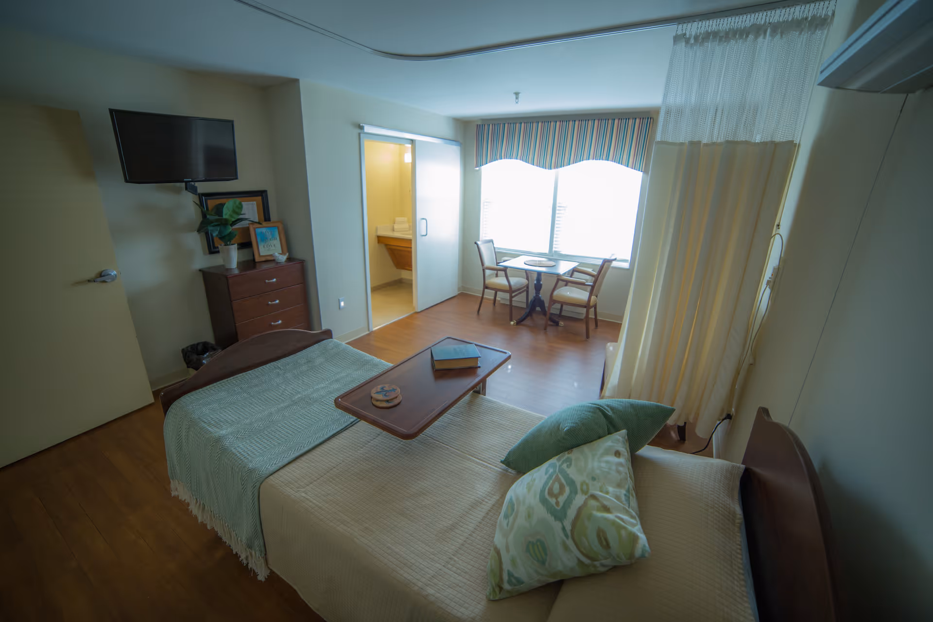 A senior living facility room with a single bed covered in beige and green bedding, a wooden overbed table with a book and coaster, a small dresser with framed pictures and a plant, a wall-mounted TV, a small table with two chairs near a large window with striped valance, and a partially visible bathroom with towels on the counter. A privacy curtain is partially drawn beside the bed.