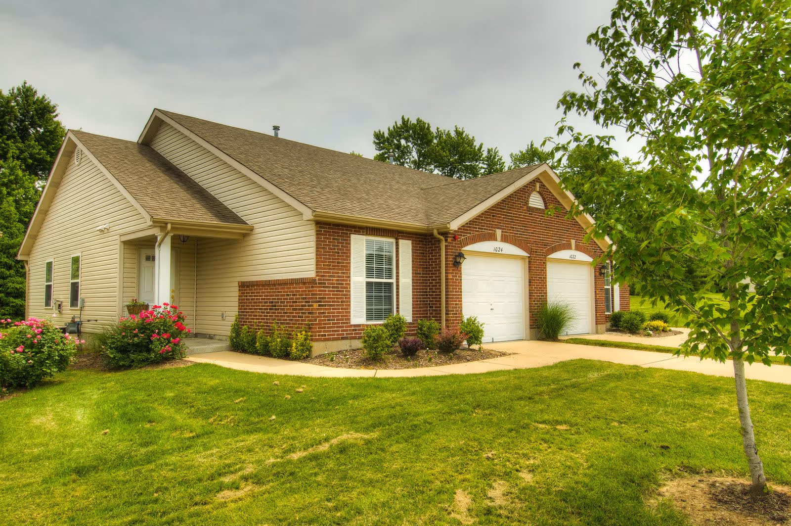 Exterior view of a single-story residential building with beige siding and red brick accents, featuring two white garage doors and a small front porch. The building is surrounded by a well-maintained lawn, bushes, and a tree on the right side.