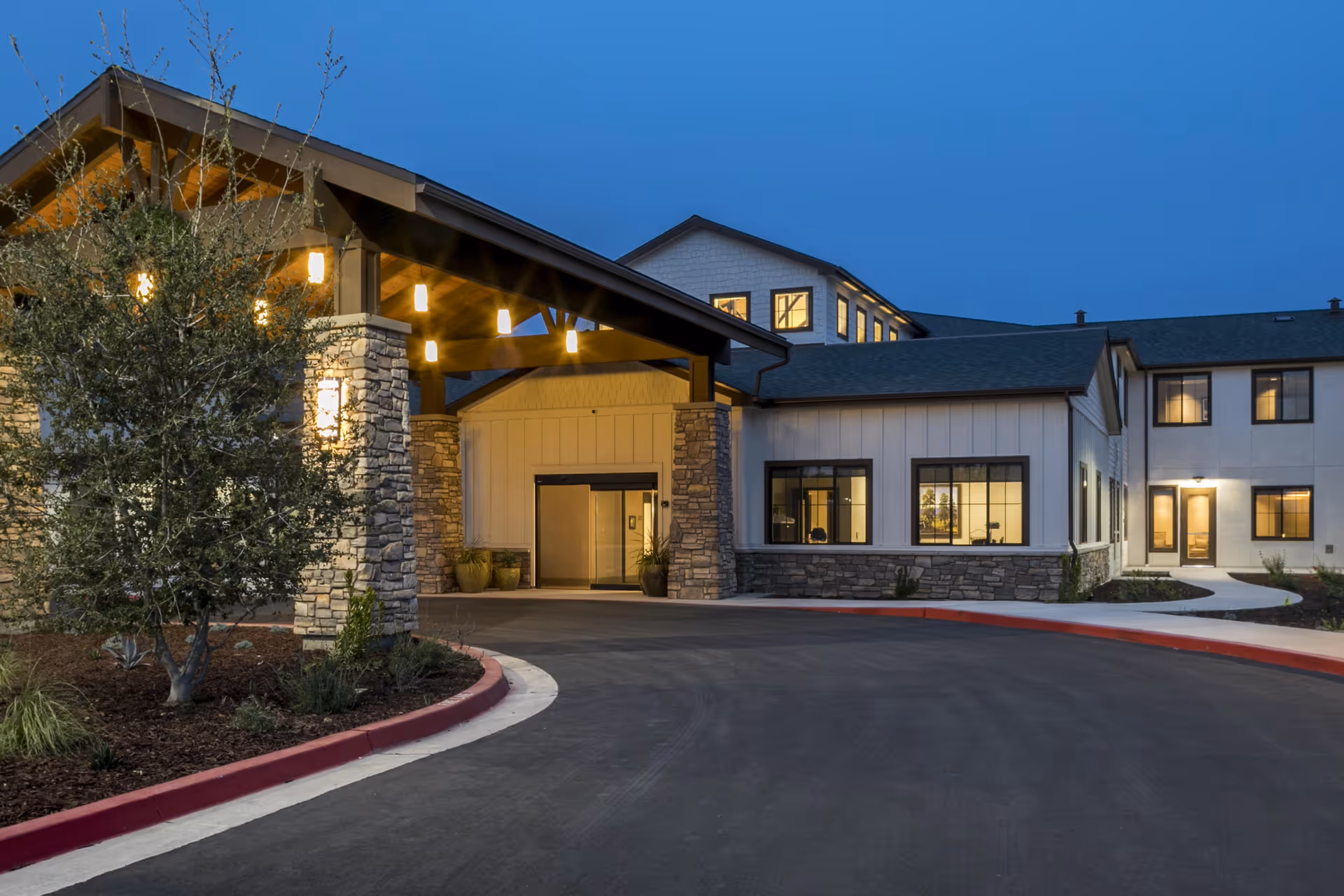 Exterior view of The Oaks at Nipomo senior living facility at dusk, showing the entrance with stone pillars, warm lighting, and a curved driveway surrounded by landscaping.