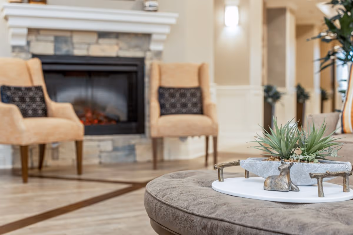A cozy senior living room area featuring a stone fireplace with a warm fire, two beige armchairs with patterned cushions, and a round ottoman with a decorative tray holding a small plant and a metallic deer figurine. The room has light-colored walls and flooring, with soft lighting and some greenery in the background.
